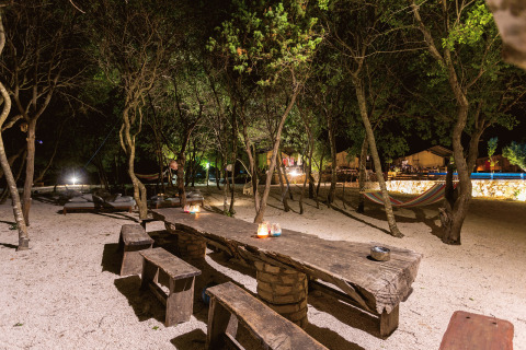 Scène nocturne au Camping Nono Ban - Safaritenten Brac avec tables en bois, bancs rustiques et hamacs entre arbres.