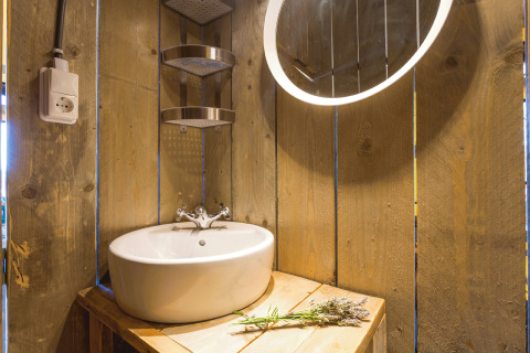 Bagno rustico con pareti in legno, lavandino rotondo, specchio illuminato e fiori in tenda safari glamping.