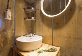 Rustic bathroom with wooden walls, round sink, illuminated mirror, and flowers in a glamping safari tent.