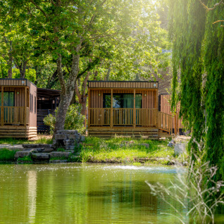 Two wooden cabins by a lakeside at Trasimeno Glamping Resort - Cottages Umbrië, surrounded by lush green trees.