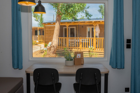 View from inside Trasimeno Glamping Resort cottage, two chairs and desk, outside a wooden terrace and tree.