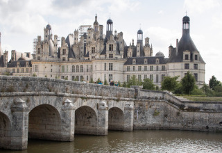 Drei Personen fahren in einem kleinen Boot auf einem Fluss, vor einem herrschaftlichen Schloss bei Huttopia Les Chateaux - Glamping Val de Loire.
