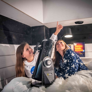 Dos personas con pijamas de estrellas usando un telescopio en una cómoda Sterrenkubussen glamping de Twente.