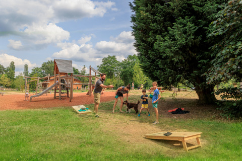 Familie spielt Cornhole und Kinder spielen auf dem Spielplatz bei Camping Huttopia De Meinweg - Glamping Limburg.