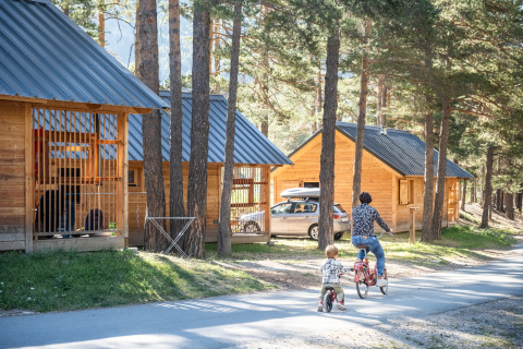 Zwei Personen fahren Fahrrad vor Holzhütten beim Glamping auf Huttopia La Clarée in den Hautes-Alpes.
