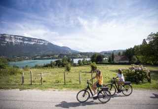 Dos personas pasean en bicicleta junto a un lago y montañas en Huttopia Lac d'Aiguebelette - Glamping Savoie.