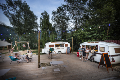 Zona glamping Huttopia Lac d'Aiguebelette con roulotte vintage, luci e ospiti che si rilassano all'aperto.
