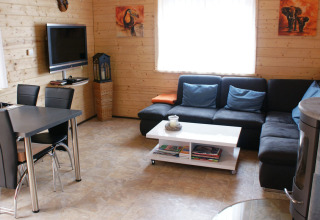 Modernes Wohnzimmer mit Holzwänden, schwarzem Sofa, Esstisch, Flachbildfernseher und bunter Deko.