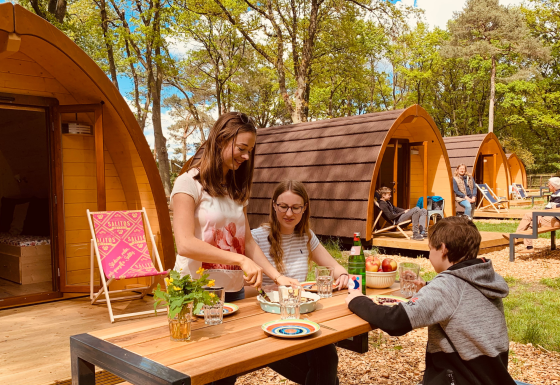 Family enjoys outdoor meal in front of wooden pods at Camping Blauer See Garbsen in forest setting.