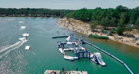 Camping Blauer See Garbsen showing inflatable water park, sandy beach, swimming guests, and green trees.