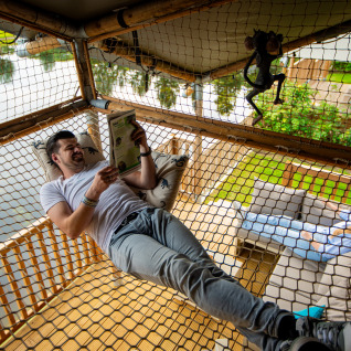 Hombre relajándose y leyendo sobre una red hamaca en Glamping De Rode Vos junto al lago en Noord-Brabant.