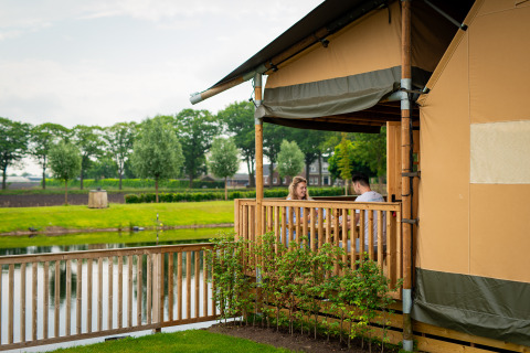 Twee personen zitten op het terras van Glamping De Rode Vos aan een vijver in Noord-Brabant, Nederland.