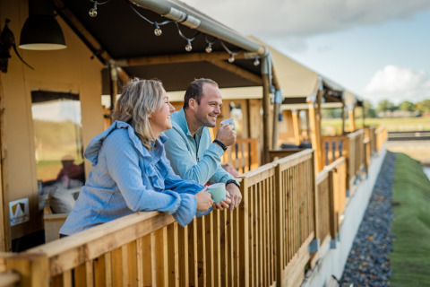Un couple boit un café sur la terrasse en bois à Glamping De Rode Vos, tentes glamping Noord-Brabant.