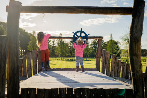 Dos niños juegan en un parque infantil de madera en Glamping De Rode Vos - Glampingtenten Noord-Brabant al sol.