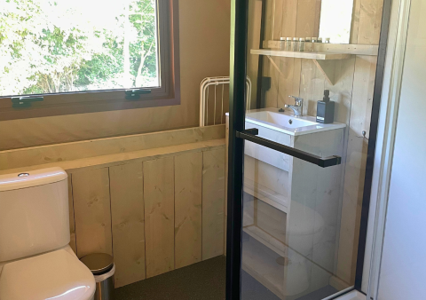 Badeværelse med træpaneler, vindue, toilet, vask og bruser i Urban Gardens Ieper glampingtelte.