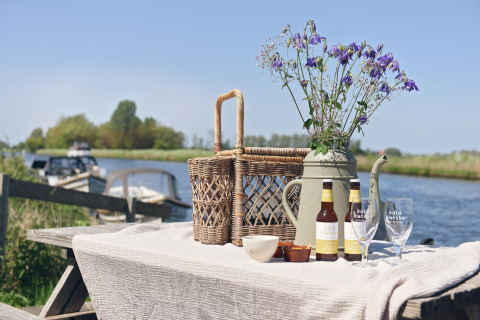 Picknicktisch mit Blumen, Korb, Bier und Gläsern am Wasser bei Smûk Recreatie, Tiny houses Friesland.