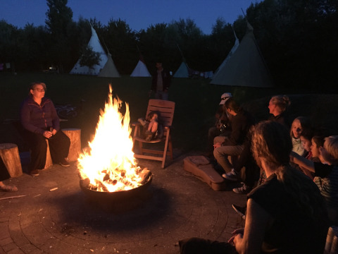 People sitting around a campfire in the evening, with teepees at Minicamping De Bokkepsprong in Zeeland.