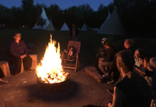 Folk sidder omkring et lejrbål ved aften, foran tipier på Minicamping De Bokkepsprong i Zeeland.