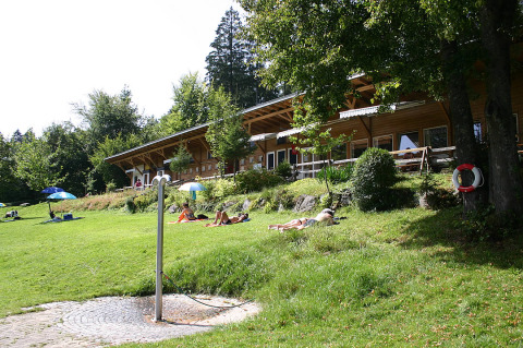 Grüne Wiese bei Camping Isny mit Sonnenbadenden und Holzgebäude, luxuriöses Glamping in Baden-Württemberg.