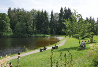 Área de camping verde junto a un lago con huéspedes, bancos y árboles en Camping Isny - Luxe glamping en Baden-Württemberg.