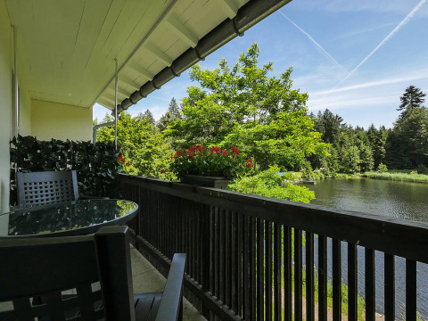 Balkon met tafel, stoelen en bloemen met uitzicht op meer en bomen bij Camping Isny - Luxe glamping Baden-Württemberg.