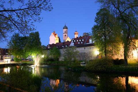 Evening view of Camping Isny - Luxe glamping Baden-Württemberg with illuminated buildings and a fountain by a pond.