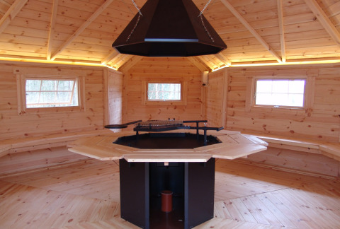 Interior de una cabaña de madera para glamping con bancos, ventanas y barbacoa central.