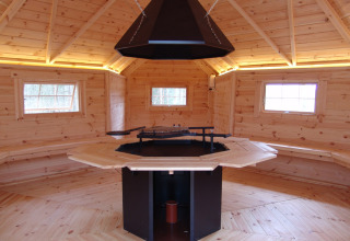 Interior of a wooden glamping hut featuring seating, windows, and a central barbecue grill setup.