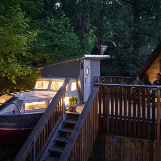 Unique glamping at Baumschiffhotel Boomschepen Beieren with a boat, terrace, and cabin surrounded by forest.