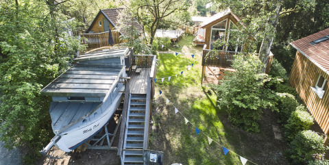 Unique glamping site with a boat cabin called Baumschiffhotel among trees, wooden lodges, and festive flags.