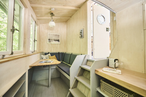 Knusse binnenkant van Baumschiffhotel glamping in Beieren met tafel, bank en trapje zichtbaar.