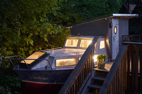 Vue du soir du Baumschiffhotel - Boomschepen Beieren glamping, cabane en forme de bateau entourée d’arbres verts.