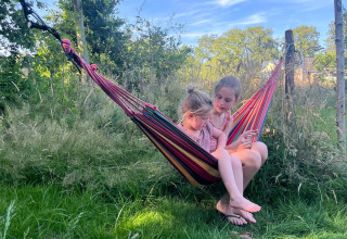 Due bambini si rilassano in un'amaca colorata sull’erba a Uitstek - Tiny Houses Drenthe glamping.