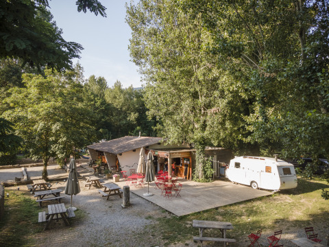 Zona de glamping al aire libre en Huttopia Millau - Glamping Aveyron, con mesas, sillas rojas y caravana blanca.