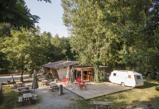Zona de glamping al aire libre en Huttopia Millau - Glamping Aveyron, con mesas, sillas rojas y caravana blanca.