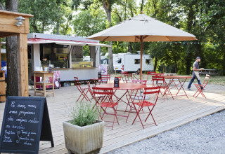 Zona de comida al aire libre con food truck y sillas rojas en Huttopia Millau - Glamping Aveyron, rodeado de árboles.