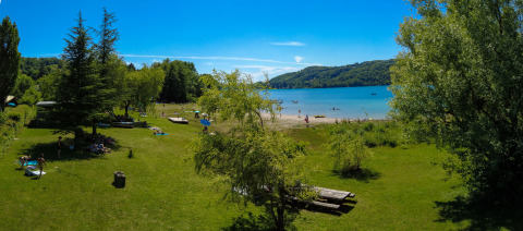 Grüner Campingplatz mit Menschen am Seeufer und Bäumen im Hintergrund bei Camping Détente et Clapotis.