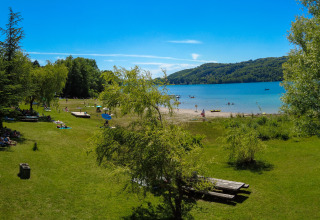 Groene kampeerplek aan een meer met mensen, bomen en heuvels bij Camping Détente et Clapotis.
