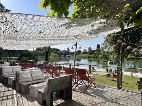 Sunny outdoor lounge with wooden benches, red chairs, and lake view at Whaka Lodge glamping in South France.