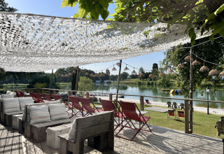 Sonnige Terrasse mit Sitzplätzen, roten Stühlen und Blick auf den See im Whaka Lodge Glamping, Südfrankreich.
