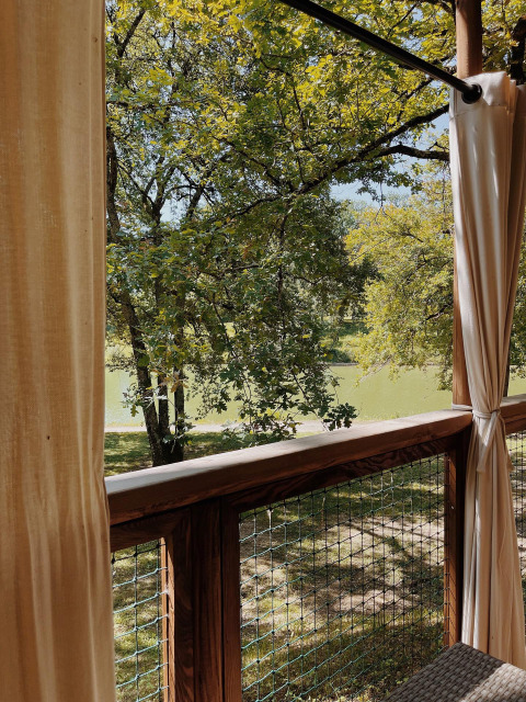Blick von einer Holzterrasse mit Vorhängen auf See und Bäume im Whaka Lodge Glamping in Südfrankreich.