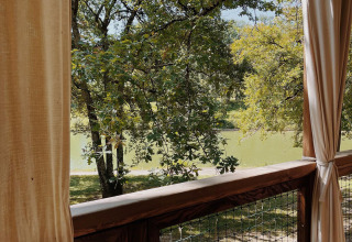 Vista desde una terraza de madera con cortinas hacia lago y árboles en Whaka Lodge glamping en el sur de Francia.