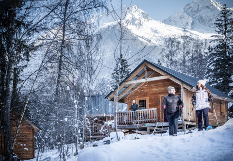 Huttopia Winter Chalets – Glamping Savoie met gasten buiten in de sneeuw en uitzicht op besneeuwde bergen.