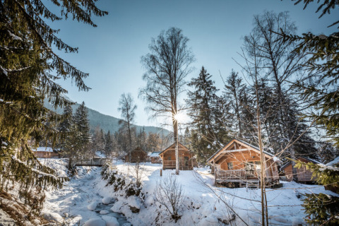 Winter glamping at Huttopia Vanoise with cozy wooden chalets, snowy forest scenery and a clear stream.