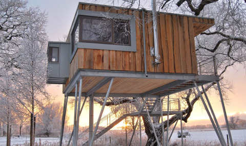Moderne Baumhütte auf Stelzen im verschneiten Winterwald bei Sonnenuntergang, Glamping Niedersachsen.
