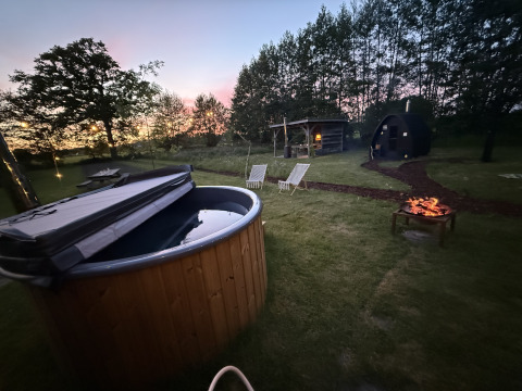 Udendørs glampingområde ved skumring med opvarmet badekar, bålplads og hyggelige træhytter i Gelderland.