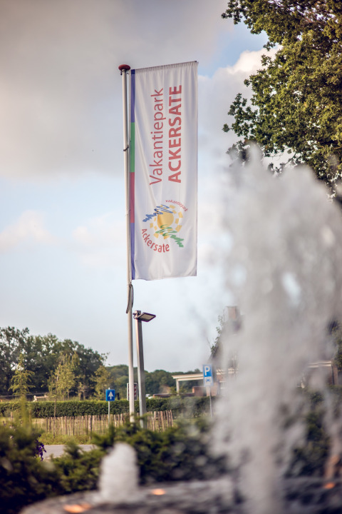 Vakantiepark Ackersate vlag wappert bij fontein, omgeven door groen, in Gelderland, Nederland, op een zonnige dag.