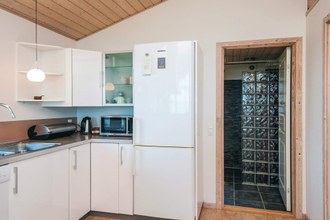 Cucina moderna con mobili bianchi, grande frigorifero e porta verso il bagno in una casa vacanze a Glesborg.
