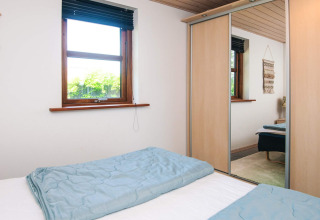 Slaapkamer met dubbel bed, blauwe dekbedden, spiegelkast en raam in vakantiewoning in Glesborg, Denemarken.