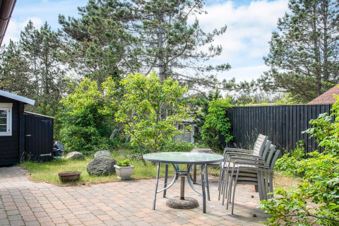 Outdoor patio with glass table and stacked chairs, surrounded by trees at a holiday home in Glesborg, Denmark.